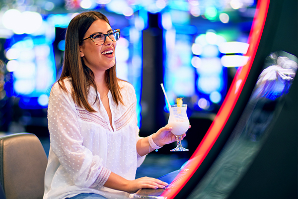 Woman In Glasses Holding A Drink Smiling While Playing At A Machine In The Casino