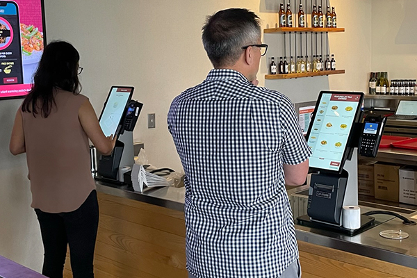 Male And Female Customers Using Countertop Kiosks To Order Meal