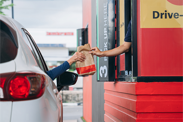 White Car In Fast Casual Restaurant Drive Thru Grabbing Brown To Go Bag With Food From Window Worker